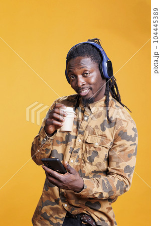 Positive smiling adult holding cup of coffee, holding smarphone scrolling to social media during lesiure time in studio. African american man searching information on internet using mobile phone Positive smiling adult holding cup of coffee, holding smarphone scrolling to social media during lesiure time in studio. African american man searching information on internet using mobile phone 104445389