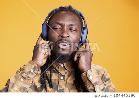 Positive stylish african american man listening radio song wearing headset, enjoying favorite playlist during free time. Cheerful adult having fun with music and dance in studio over yellow background 104445390