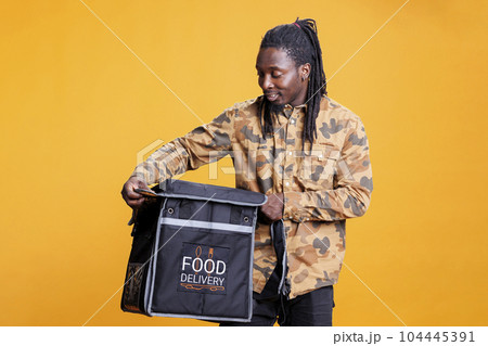 Deliveryman holding thermal backpack carrying takeaway food, delivering customers orders during lunch time. African american restaurant courier standing in studio with yellow background. Deliveryman holding thermal backpack carrying takeaway food, delivering customers orders during lunch time. African american restaurant courier standing in studio with yellow background. 104445391