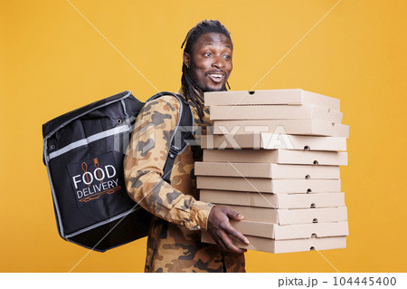 Pizzeria delivery employee holding cardboard stack full with pizza, delivering takeaway food order to client during lunch time. African american deliveryman carrying thermal backpack in studio 104445400