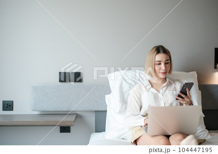 Cheerful asian woman looking a message from a Phone Using Laptop Sitting In Bed At Home. Cheerful asian woman looking a message from a Phone Using Laptop Sitting In Bed At Home. 104447195