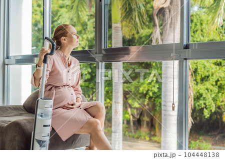 Young pregnant woman enjoys cleaning her house. Easy cleaning with a wireless vacuum cleaner. Pregnant woman cleaning floor with handheld vacuum cleaner. Modern easy cleaning 104448138