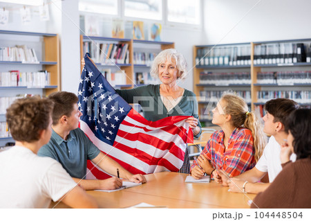 Mature female teacher shows the flag of United States of America to teenagers and talks about this country during lesson at library school 104448504