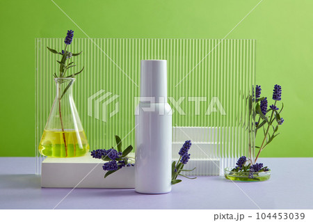 Front view of white bottle unbranded displayed on green background with lavender, white podiums and acrylic sheet. Lavender oil could be a gentle way to treat acne due to its ability to kill bacteria. Front view of white bottle unbranded displayed on green background with lavender, white podiums and acrylic sheet. Lavender oil could be a gentle way to treat acne due to its ability to kill bacteria. 104453039