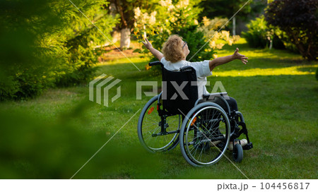 Rear view of an elderly woman spread her arms to the sides while sitting in a wheelchair on a walk outdoors. Rear view of an elderly woman spread her arms to the sides while sitting in a wheelchair on a walk outdoors. 104456817
