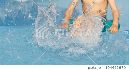 Happy kid splashing water in blue swimming pool on sunny day at summer vacation. Little boy child siting on the edge of the pool, his legs dipping in the water. Copy space 104461670