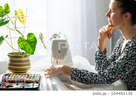 Woman thinking, dreaming and sews tulle on an electric sewing machine in a white modern interior of a house with large windows, house plants. Comfort in house, housewife's hobby 104462136