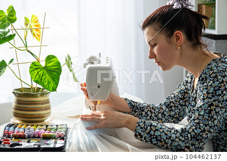 A woman sews tulle on an electric sewing machine in a white modern interior of a house with large windows, house plants. Comfort in the house, a housewife's hobby 104462137