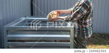 A woman in a plaid shirt pours food waste from a bowl into a compost heap of potato and carrot peelings. Compost box made of metal, eco-friendly fertilizer for the garden A woman in a plaid shirt pours food waste from a bowl into a compost heap of potato and carrot peelings. Compost box made of metal, eco-friendly fertilizer for the garden 104462303