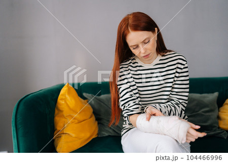 Front view of suffering from pain injured young woman with broken right hand wrapped in white gypsum bandage sitting on couch closed eyes. Sad redhead female with broken arm from accident at home. 104466996