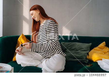 Positive injured young woman with broken right arm wrapped in white gypsum bandage applying cosmetic on face with professional makeup brush, looking to mirror sitting on sofa at home. 104466997