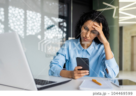 Shocked and upset business woman working inside office at workplace, depressed hispanic woman reading bad news using app on smartphone, holding phone. Shocked and upset business woman working inside office at workplace, depressed hispanic woman reading bad news using app on smartphone, holding phone. 104469599