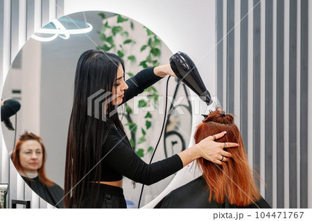 Woman hairdresser works in beauty salon and does hair styling with hairdryer. Beautiful oriental 104471767