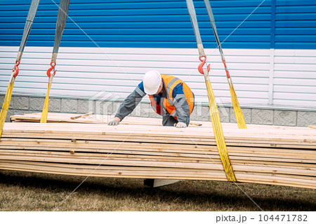 Slinger unloads wooden boards on ground. Worker in helmet and high-visibility vest supervises Slinger unloads wooden boards on ground. Worker in helmet and high-visibility vest supervises 104471782