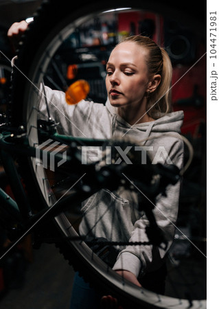 Vertical front view of blonde cycling mechanic female repairing and fixing mountain bicycle standing on bike rack in repair workshop with dark interior. Concept of professional bicycle maintenance. 104471981