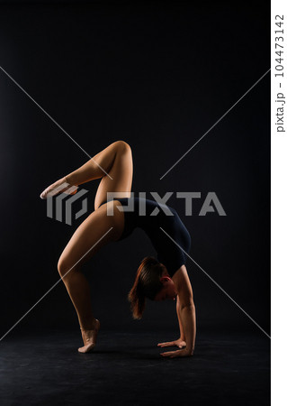 A girl gymnast in a swimsuit does tricks on a white and dark background. Front view A girl gymnast in a swimsuit does tricks on a white and dark background. Front view 104473142