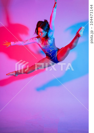 A girl gymnast in a swimsuit does tricks on a white and dark background. Front view A girl gymnast in a swimsuit does tricks on a white and dark background. Front view 104473144