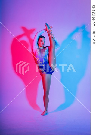 A girl gymnast in a swimsuit does tricks on a white and dark background. Front view 104473145
