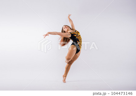 A girl gymnast in a swimsuit does tricks on a white and dark background. Front view 104473146