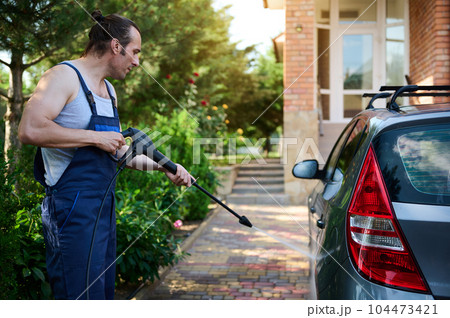 Caucasian handsome muscular man 40 years old, wearing a blue work uniform, directing a spray wand with water jet while washing car under high pressure outdoors. Car cleaning concept. People. Transport Caucasian handsome muscular man 40 years old, wearing a blue work uniform, directing a spray wand with water jet while washing car under high pressure outdoors. Car cleaning concept. People. Transport 104473421