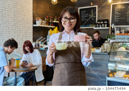 Portrait of smiling female coffee shop worker holding two cups of coffee in hands Portrait of smiling female coffee shop worker holding two cups of coffee in hands 104475494