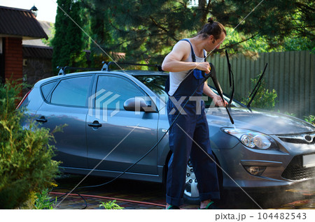 Young car service worker in blue uniform, directs sprayer at headlight, manually washes car under high pressure water jet. The concept of people, technology and transport. Water high pressure cleaner 104482543