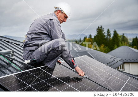 Man technician mounting photovoltaic solar panels on roof of house. Engineer in helmet installing solar module system with help of hex key. Concept of alternative, renewable energy. 104485300