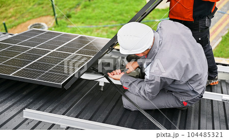 Mounters connecting cables while installing photovoltaic solar panels on roof of house. Workers mounting of PV solar modules on rooftop. Concept of alternative and renewable energy. 104485321
