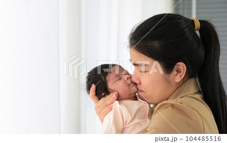 Asian young mom kissing her cute newborn little baby girl sleeping on mom arms near window at home 104485516