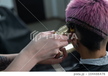 The hairdresser shaves the temple of a female client. Rear view of a woman with short pink hair in a barbershop. 104486476