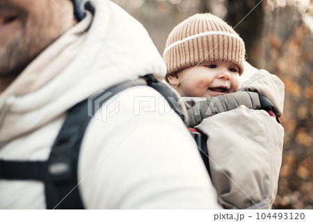 Sporty father carrying his infant son wearing winter jumpsuit and cap in backpack carrier hiking in autumn forest Sporty father carrying his infant son wearing winter jumpsuit and cap in backpack carrier hiking in autumn forest 104493120