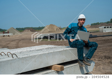 Caucasian male builder in hardhat sits on floor slabs and uses laptop at construction site.  104504528