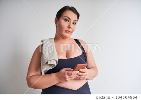 Sporty fat woman holding smartphone and poses camera in white wall 104504964