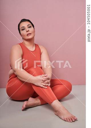 Confident woman in sportswear looking at camera and sitting over pink studio wall 104505001