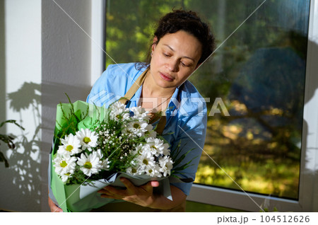 Charming ethnic woman florist, decorator holds a beautiful bouquet of white daisies and chamomile flowers, arranged in a floral studio design for holidays or festive life events. Arrangement and gifts Charming ethnic woman florist, decorator holds a beautiful bouquet of white daisies and chamomile flowers, arranged in a floral studio design for holidays or festive life events. Arrangement and gifts 104512626
