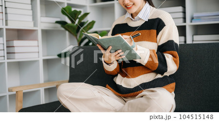 Young Asian woman listening music from headphones and writing note for her work idea in diary book.She in living room on the sofa at home Young Asian woman listening music from headphones and writing note for her work idea in diary book.She in living room on the sofa at home 104515148