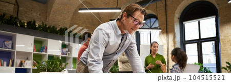 Successful businessman standing in the modern coworking space, looking at laptop screen and thinking about something while his young colleagues talking on the background 104515521