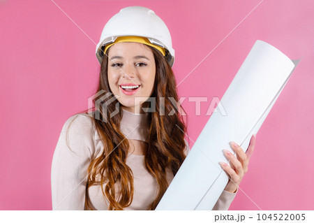 Portrait of a happy female architect builder in the hard hat holding layout plan on the pink background. Portrait of a happy female architect builder in the hard hat holding layout plan on the pink background. 104522005