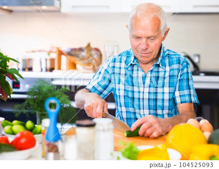 elderly man cuts vegetables for salad at the table in the kitchen 104525093