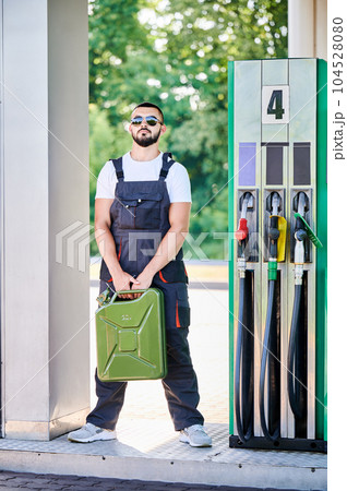Professional worker holding canister in hands. Confident operator in sunglasses and overalls waiting for customers. Male worker standing on background of gas station with cistern in hands. 104528080