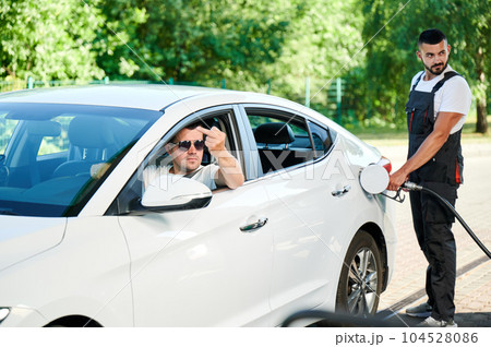 Rude driver showing middle finger to other car on gas station. Angry man in expensive car showing an indecent gesture through window. Gas station operator smiling at rude client. 104528086