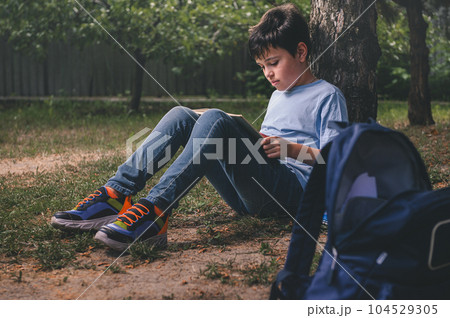 Handsome diligent teenage schoolboy in casual denim clothes, sitting under a tree in the park, focused on doing homework Handsome diligent teenage schoolboy in casual denim clothes, sitting under a tree in the park, focused on doing homework 104529305