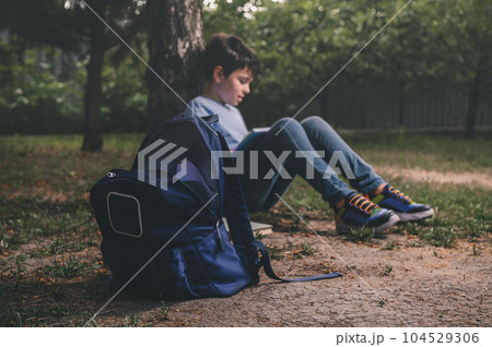Focus on backpack over smart teenage schoolboy relaxing after class, doing homework in the park, sitting under tree Focus on backpack over smart teenage schoolboy relaxing after class, doing homework in the park, sitting under tree 104529306