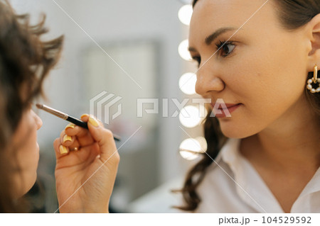 Closeup of female makeup artist applying cosmetic tonal foundation for face sculpting of pretty young woman in beauty studio. Creating creative image for photoshoot. Make-up artist corrects make-up. 104529592