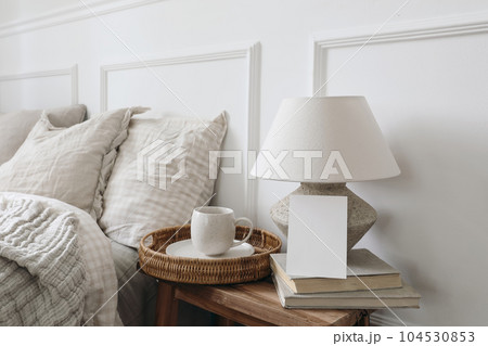 Closeup of blank greeting card, invitation mockup. Breakfast in bed concept. Cup of coffee, table lamp with linen shade, vintage wooden night stand. Checkered beige pillows, blurred background. 104530853