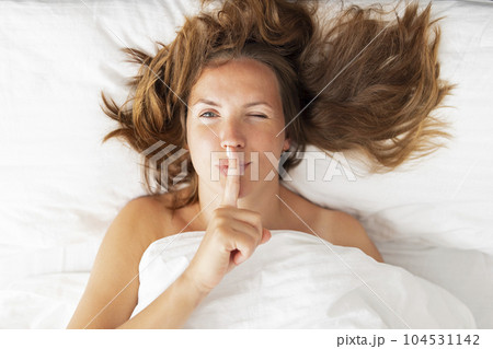 Portrait of a woman lying in bed covered with a blanket holding finger on lips 104531142