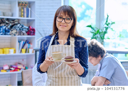 Portrait of female in apron holding fresh cup of coffee and cake on plate in coffee shop 104531776