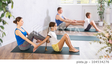 Middle-aged mother and father helping to preteen daughter and son to warming up their back before family yoga training together in gym fitness center 104534276