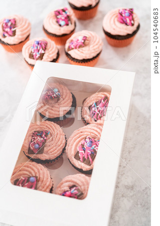 Packaging freshly baked chocolate strawberry cupcakes garnished with gourmet mini pink chocolates into a white paper cupcake box. 104540683
