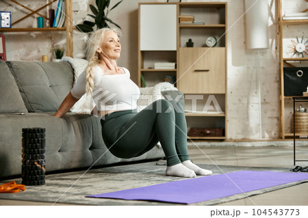 Smiling, beautiful, senior woman training at home in living room, doing exercises with couch. Strong fit body 104543773